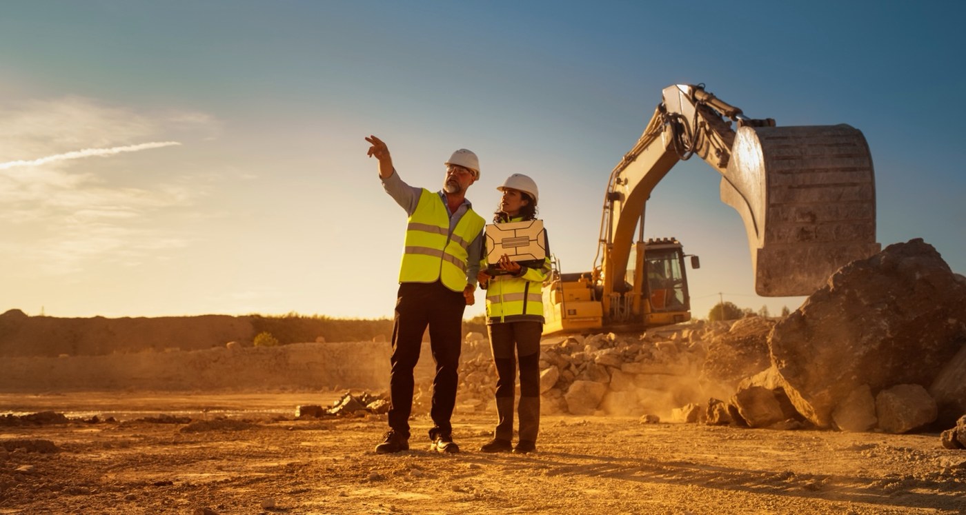 Two construction workers in hi-vis vests and hard hats on an earthwork site at golden hour, an excavator working behind them
