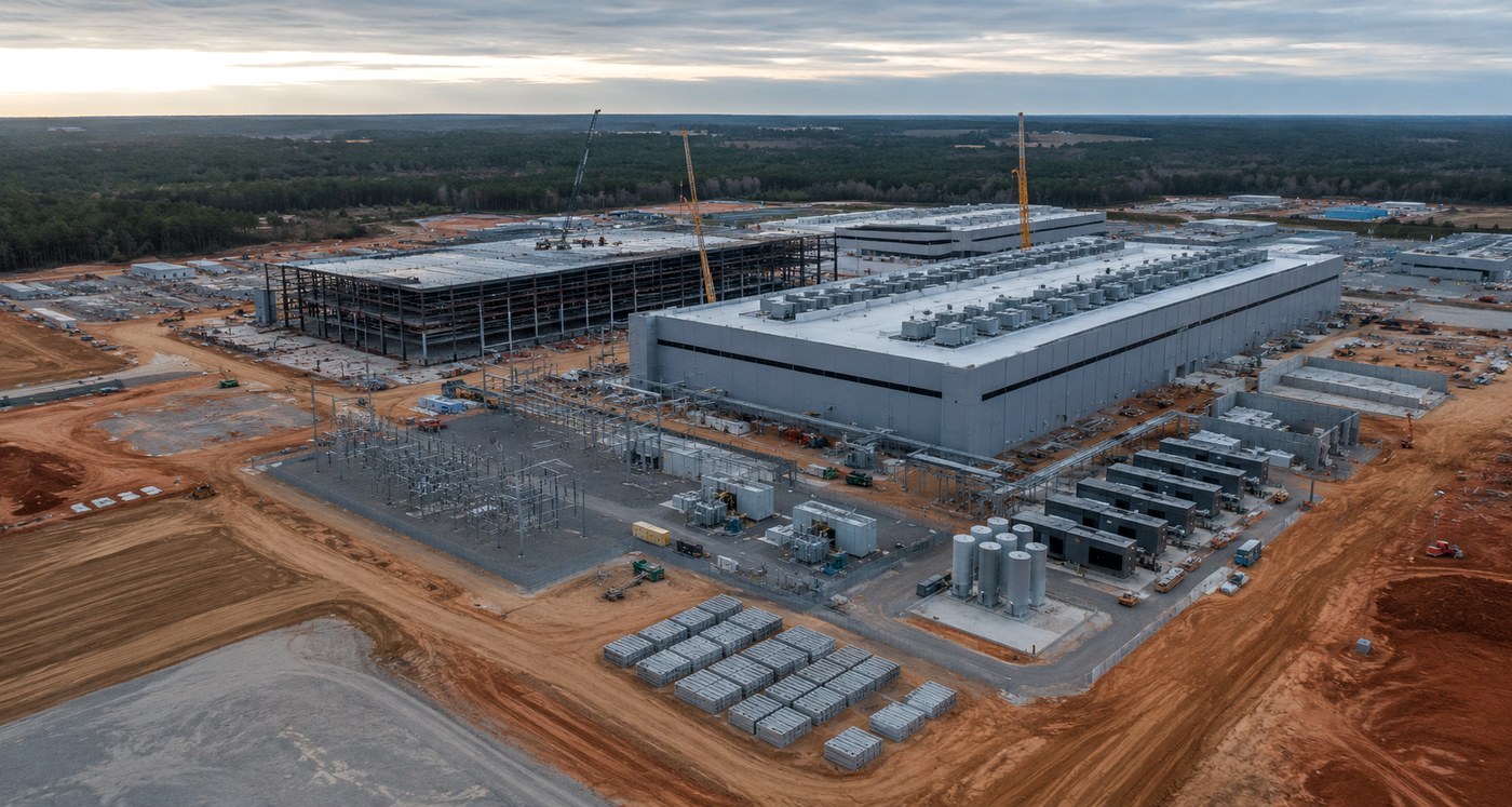 Aerial view of a hyperscale data center campus under construction on cleared red-clay land, with structural steel, rooftop cooling units, an electrical substation and tower cranes