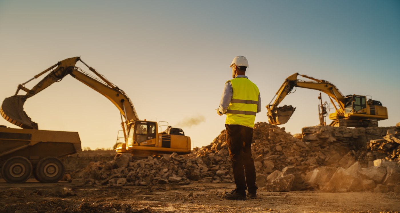 Construction worker in a hi-vis vest and hard hat overlooking an active earthwork site at golden hour, excavators loading rock behind him