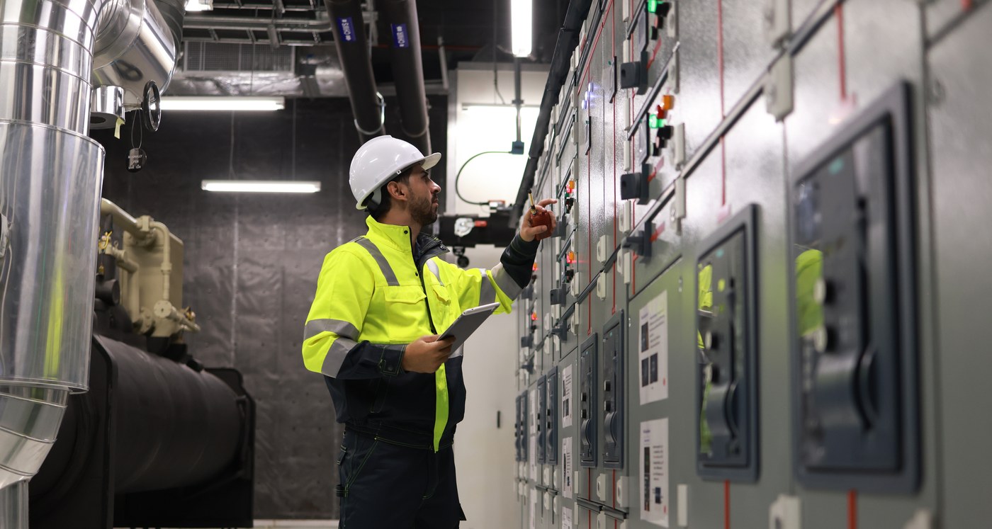 An electrician in a hi-vis vest inspects a switchgear lineup in a dark electrical room