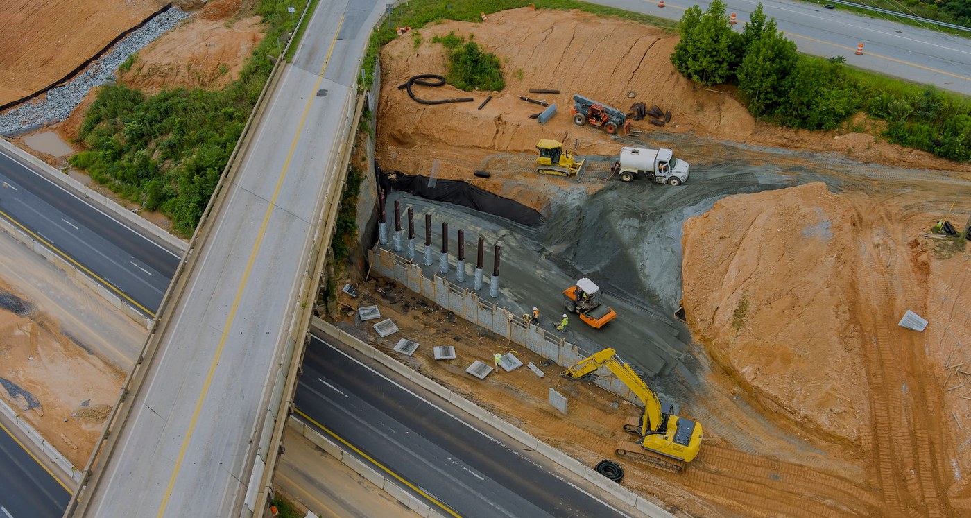 Aerial view of an interstate reconstruction: a bridge deck under construction beside live traffic, excavators and a dump truck working the graded earth below