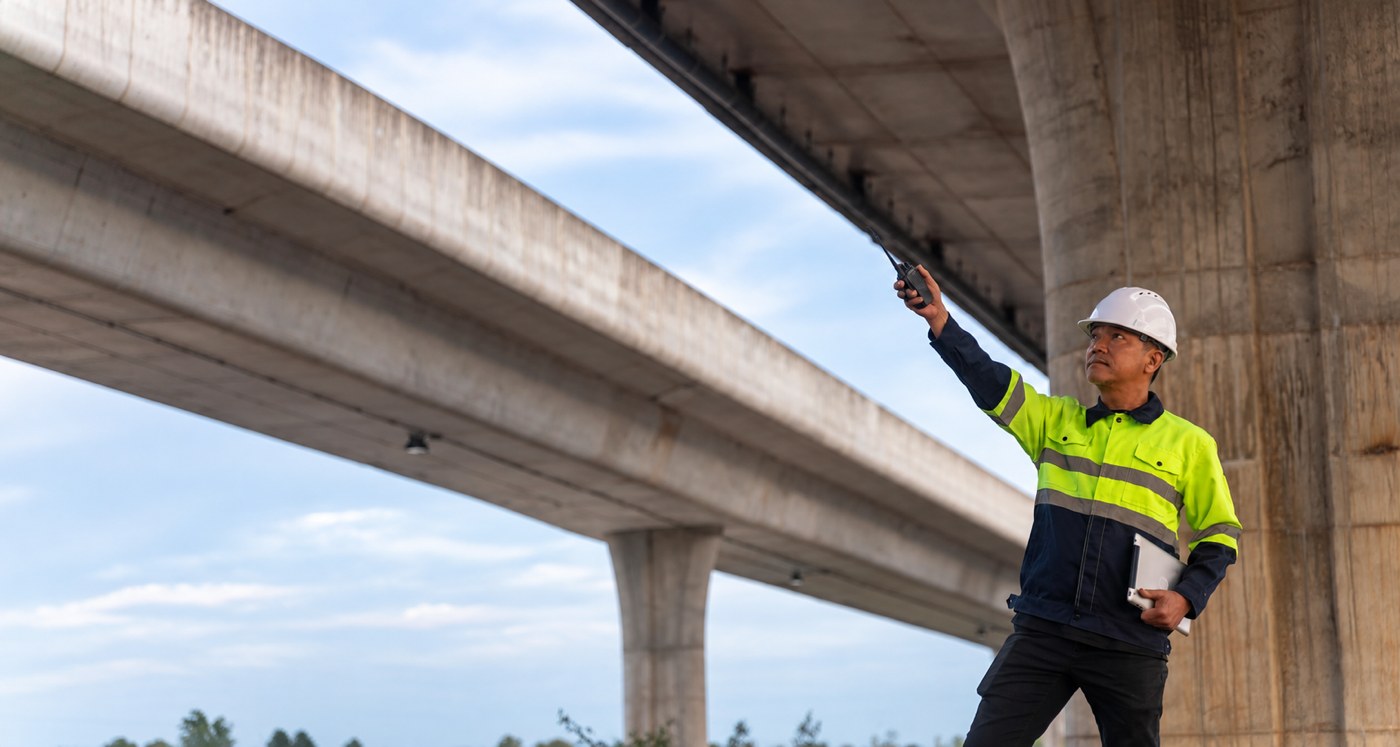 A superintendent in a white hardhat and hi-vis gear points up at bridge girders, radio in hand