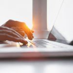 A person typing on a laptop with the sun shining in the background.